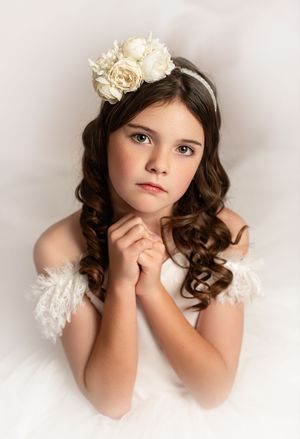 Portrait of a young subject in white dress with curled brown hair wearing a floral headpiece against neutral background.