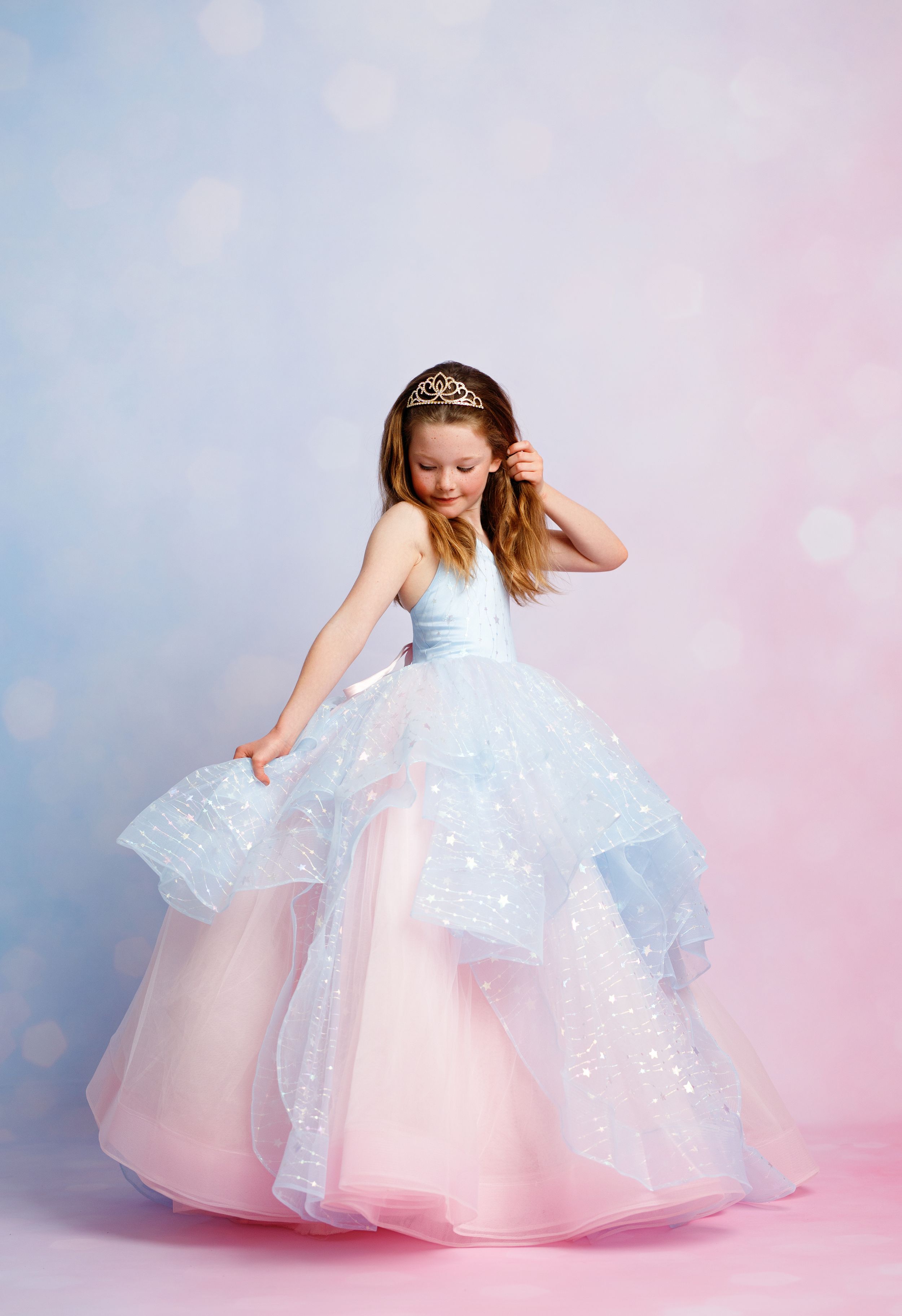 Young girl in a sparkling blue and pink tiered ball gown with a tiara, posing gracefully against a soft bokeh background.