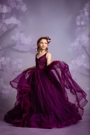 Young child in flowing deep purple ball gown poses against dreamy purple background with soft shadows.