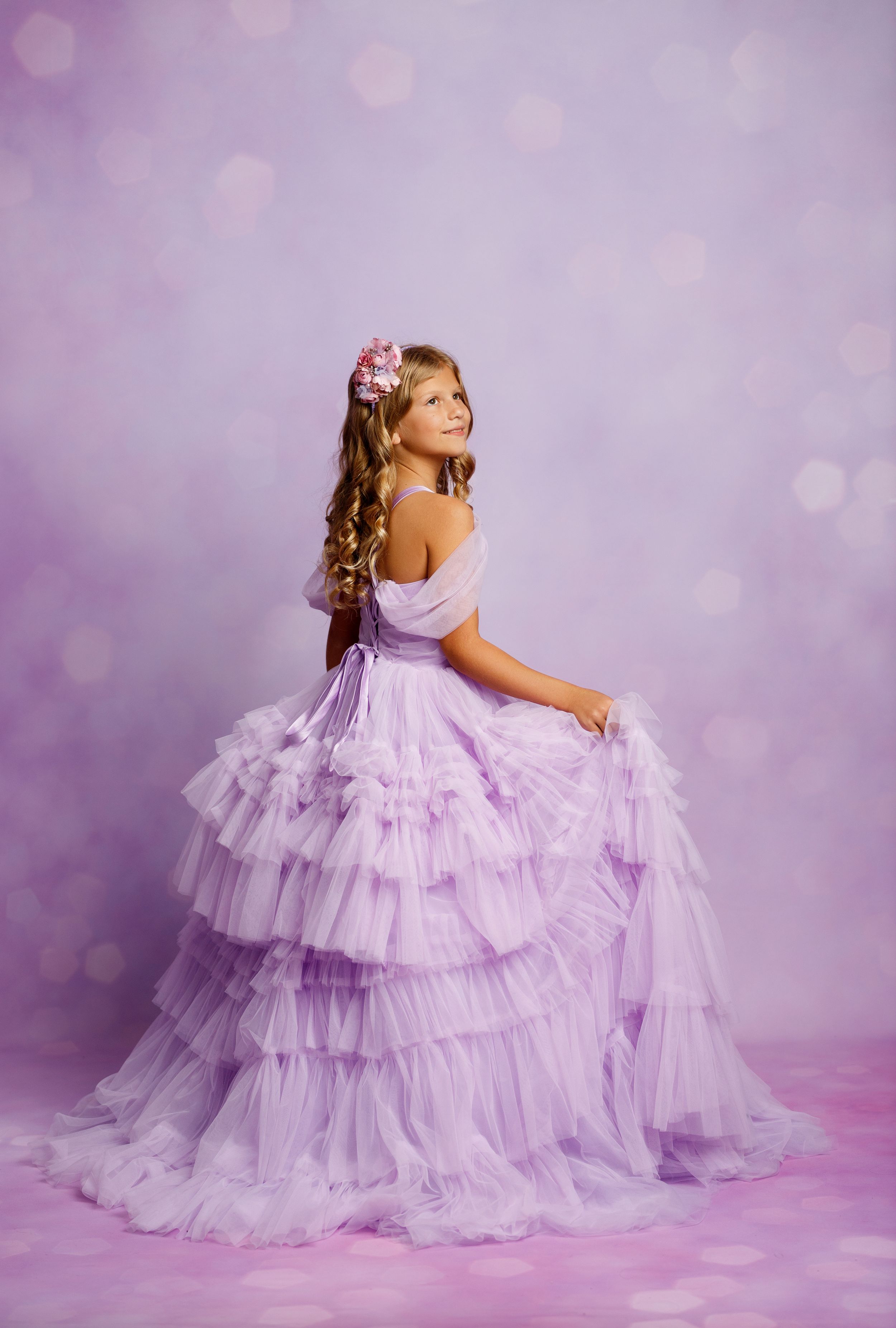 A young girl in a layered lavender tulle ball gown with floral hair accessory poses gracefully against a purple bokeh background.
