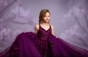A person in an elegant plum-colored ball gown with a flowing tulle skirt poses against a dreamy purple background.