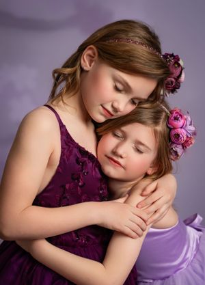 Two young girls in purple dresses embrace tenderly while adorned with pink flowers in their hair against a soft purple backdrop.