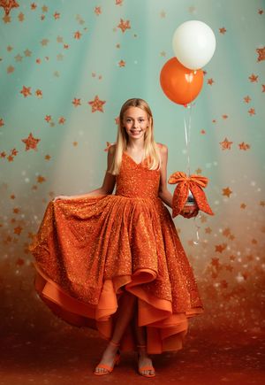 A bright orange sparkly dress twirls against a teal background with glittering star decorations and white balloons.