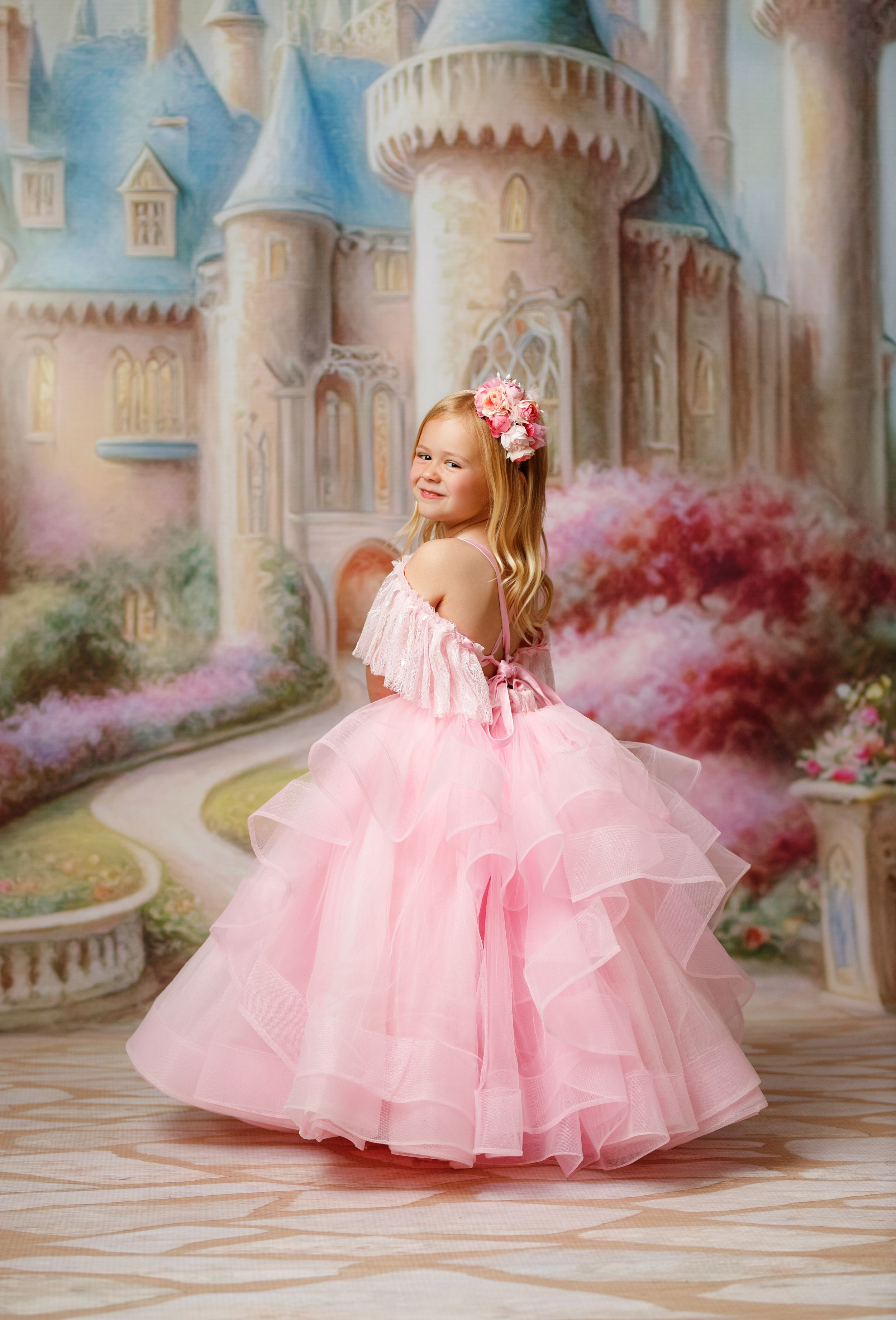 Young girl in pink ruffled ball gown with floral hair accessory poses before a fairy tale castle backdrop.