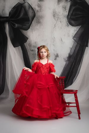A young child in a flowing red formal gown poses beside a red wooden chair against a dramatic black and white backdrop.