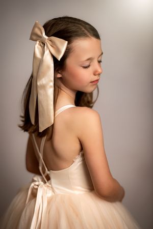 A young dancer in a cream tulle dress and satin hair bow poses elegantly with back turned to camera.