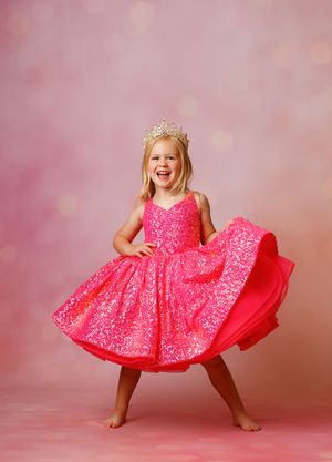 Young girl in pink sequin dress and gold crown twirls joyfully against a soft pink bokeh background.