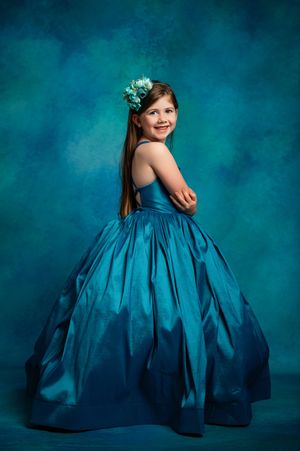 Young figure in flowing teal ball gown twirls gracefully against a deep turquoise studio backdrop.
