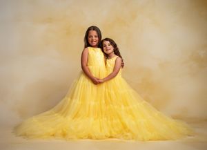 Two figures in matching yellow tulle gowns pose together against a warm beige backdrop for a portrait.