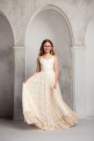 Young girl in elegant cream lace gown posing in front of arched columns in a studio setting.