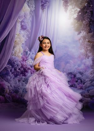 Young girl in lavender ruffle gown poses before a dreamy purple floral backdrop with flowing curtains.
