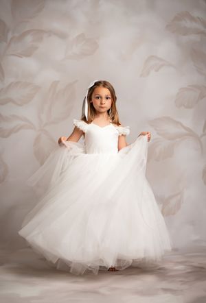 A young person in a flowing white ball gown with tulle skirt poses against a light floral background.