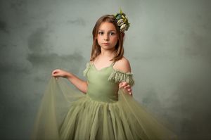 A child in a sage green tulle dress with off-shoulder sleeves and floral headpiece poses against a textured backdrop.