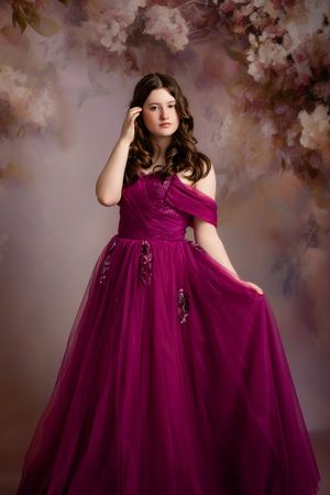 A model in a luxurious burgundy tulle gown poses against a dreamy floral backdrop in a romantic portrait session.