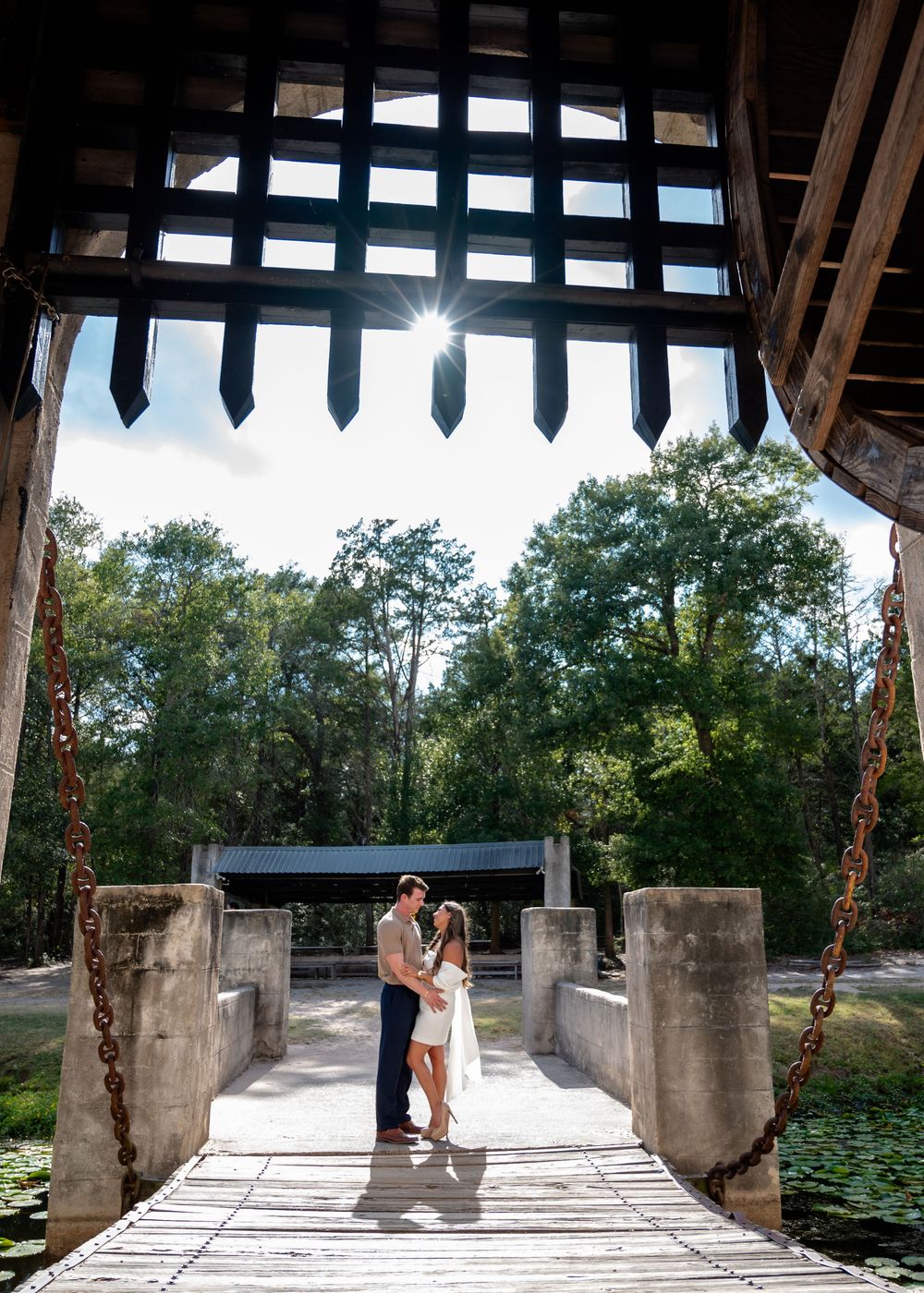 Romantic couple kissing on wooden drawbridge surrounded by lush green trees and sunlight.