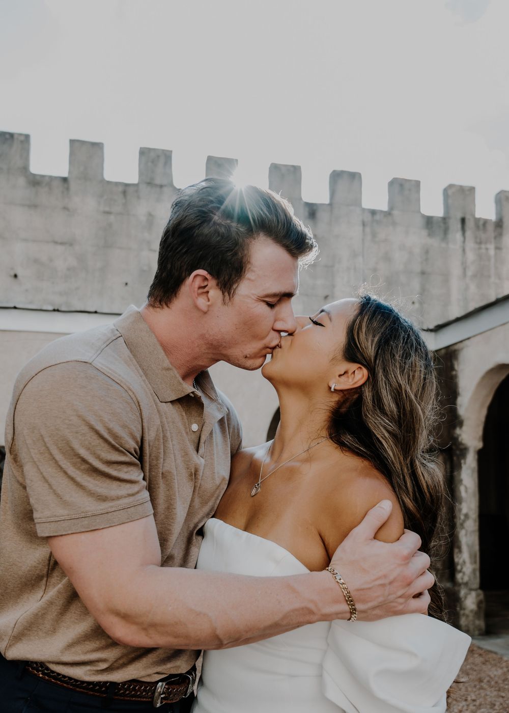 Couple sharing passionate kiss in front of castle wall with golden sunflare overhead.