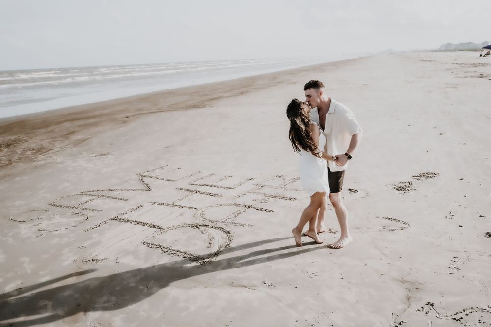Couple kissing on a sandy beach with words written in the sand, romantic engagement photo by the ocean.