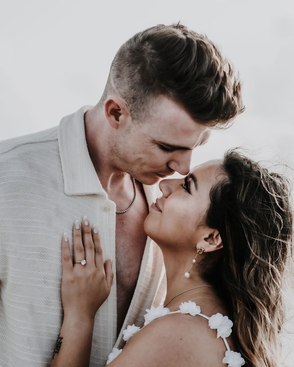 A romantic couple sharing an intimate moment, almost kissing, dressed in white outfits, with an engagement ring visible.