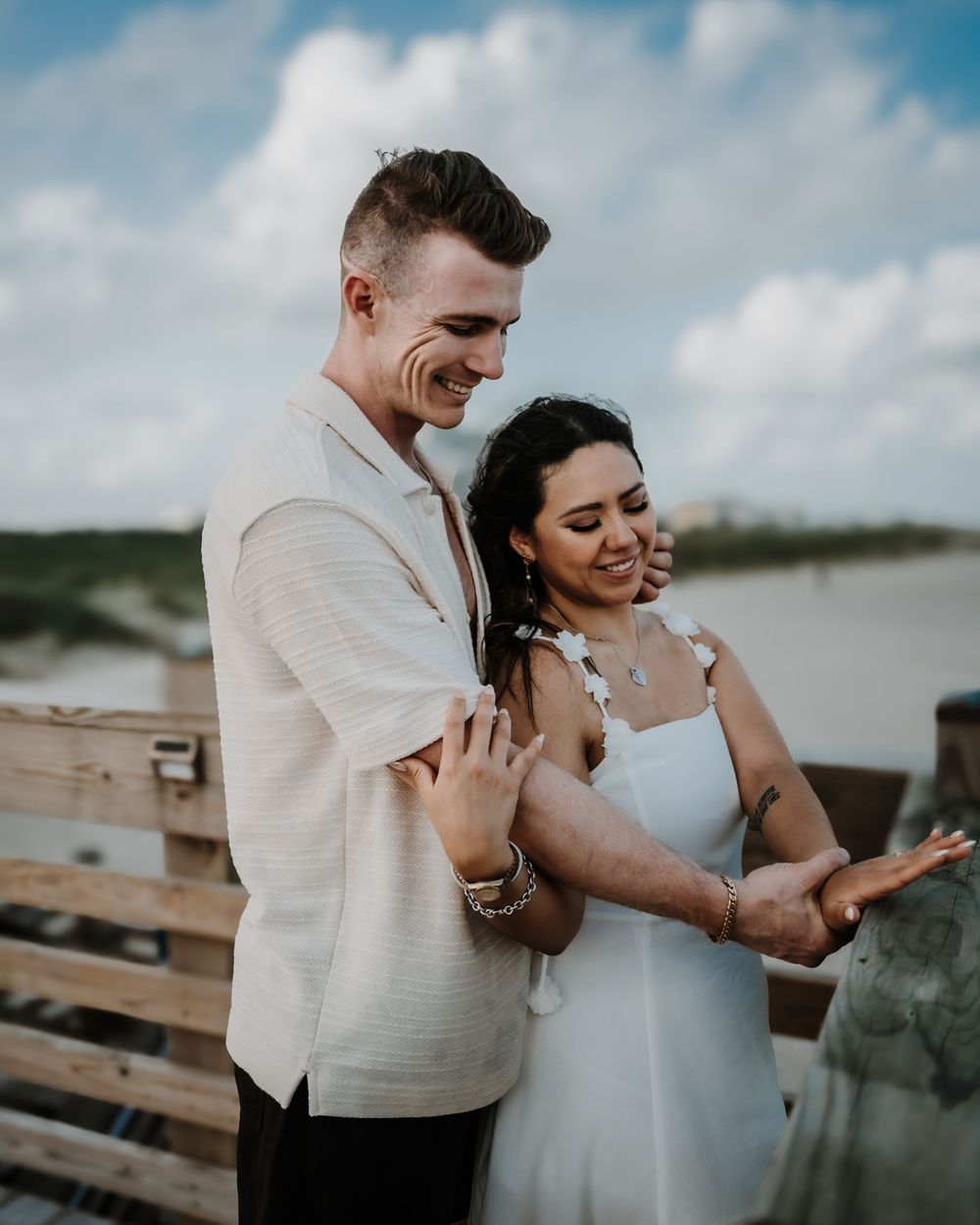 A smiling couple embraces on a beach boardwalk, woman in white dress and man in cream shirt, with ocean and clouds behind.