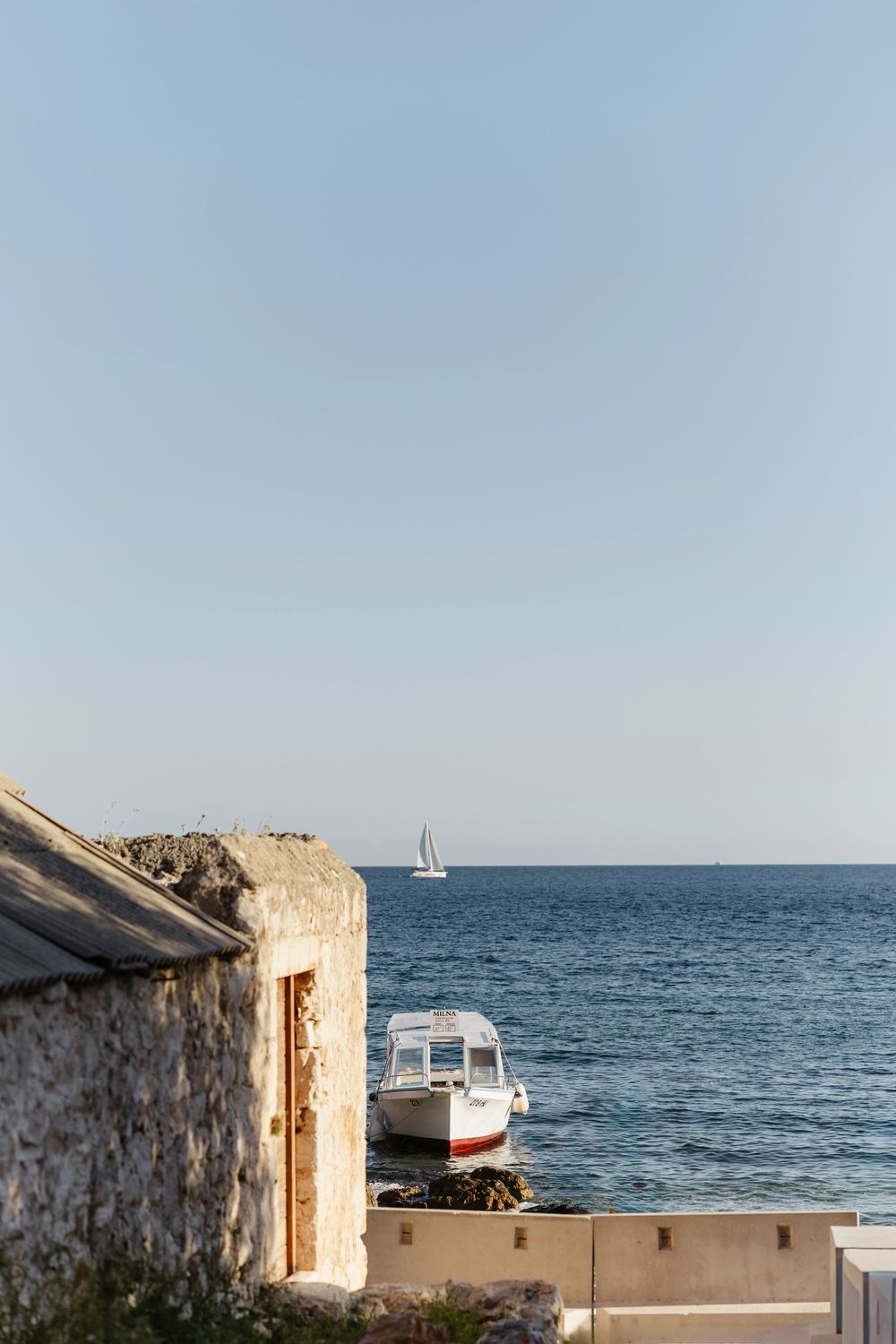 A serene coastal scene with a white boat and sailboat visible on the Mediterranean horizon near stone buildings. Hvar, Croatia
