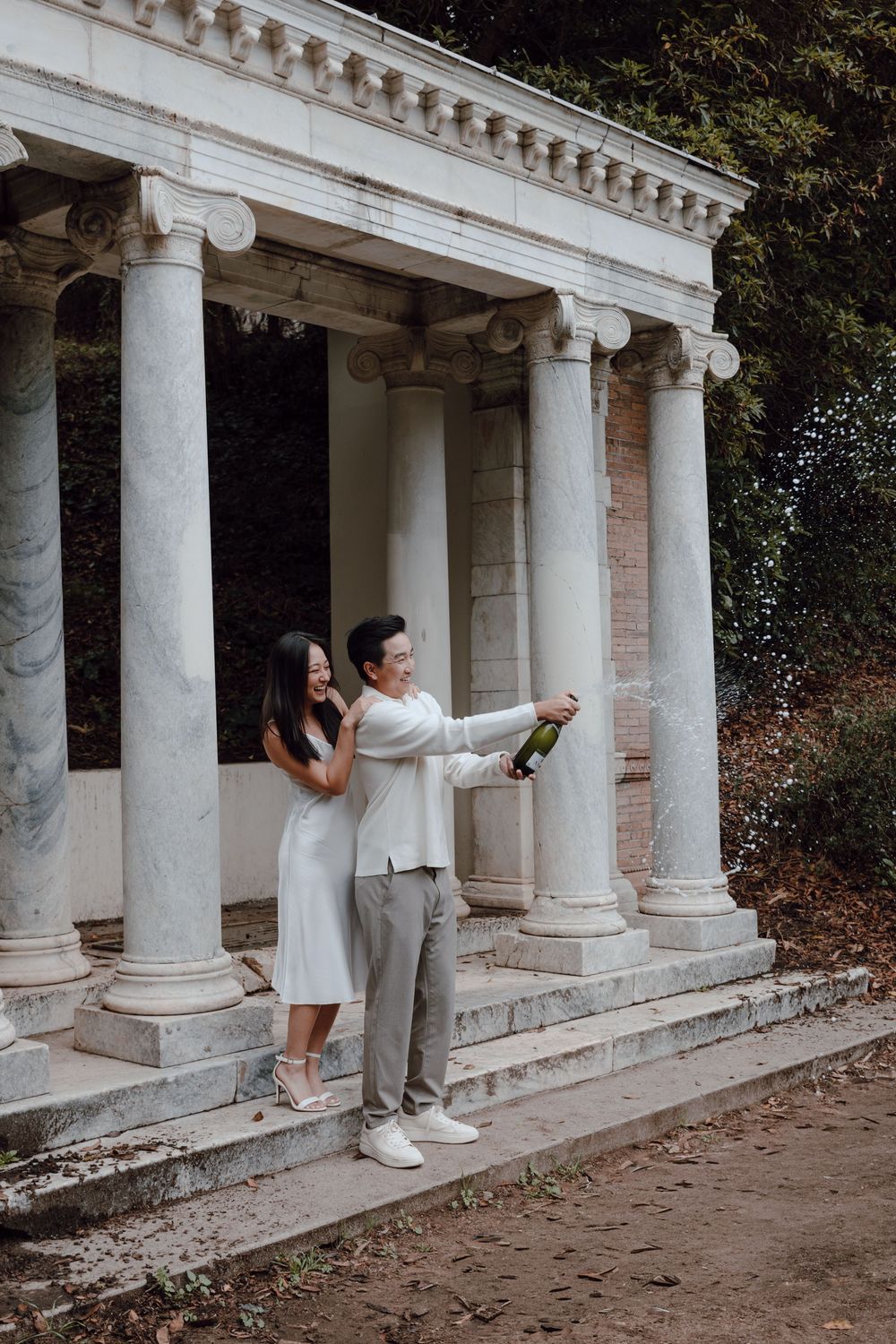 A couple poses playfully behind white marble columns of a historic colonnade in a romantic outdoor photoshoot.
