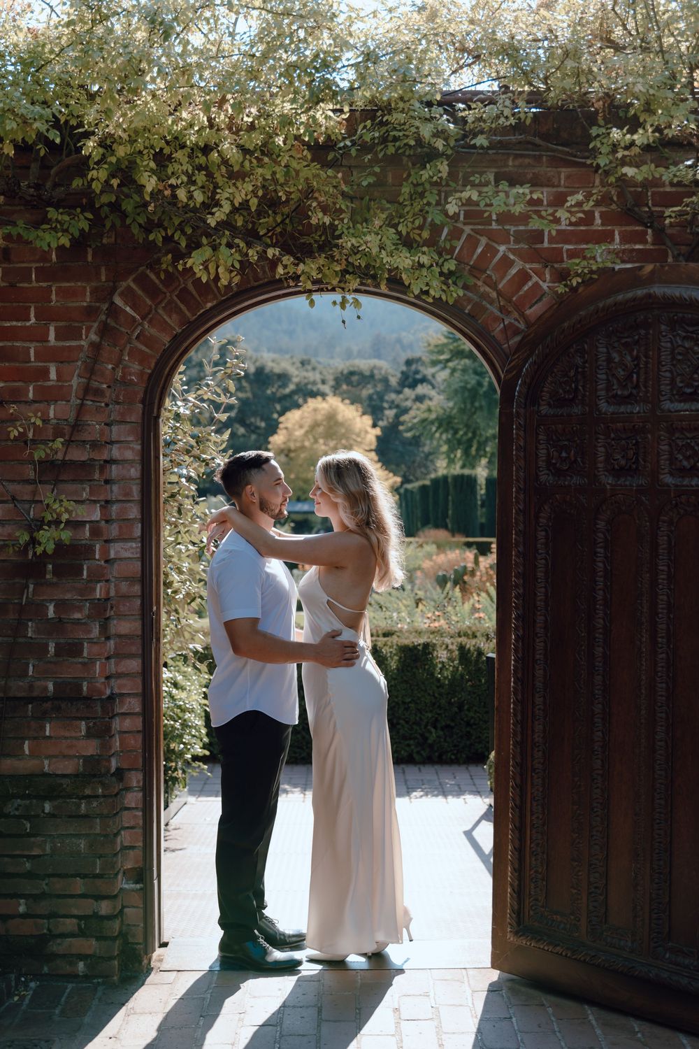 A romantic couple dances together under a brick archway surrounded by blooming white flowers on a sunny day.