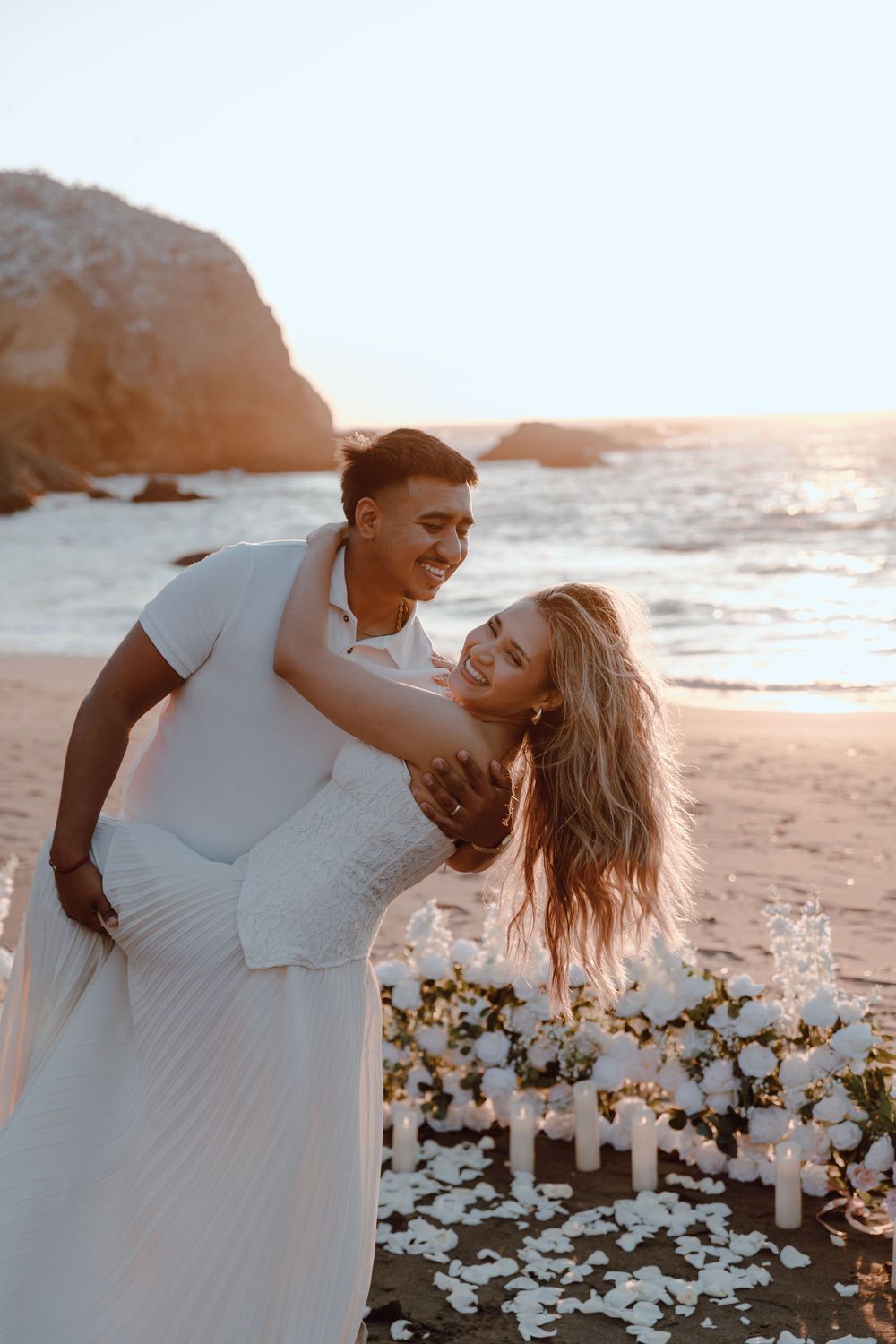 A romantic couple embraces on a sandy beach at sunset with white flowers scattered along the shoreline.