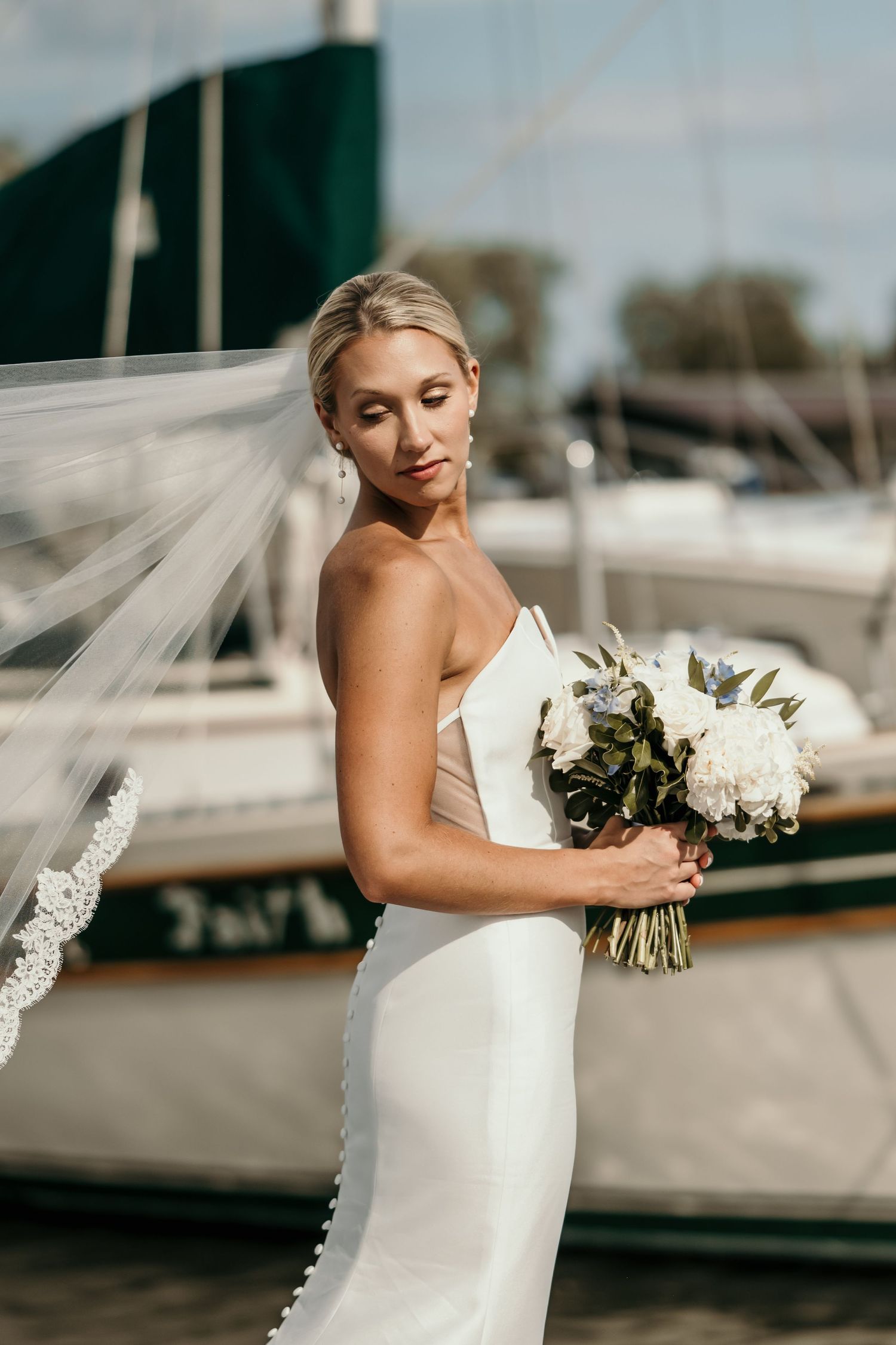 Elegant bride in white dress holding bouquet near boats at marina.