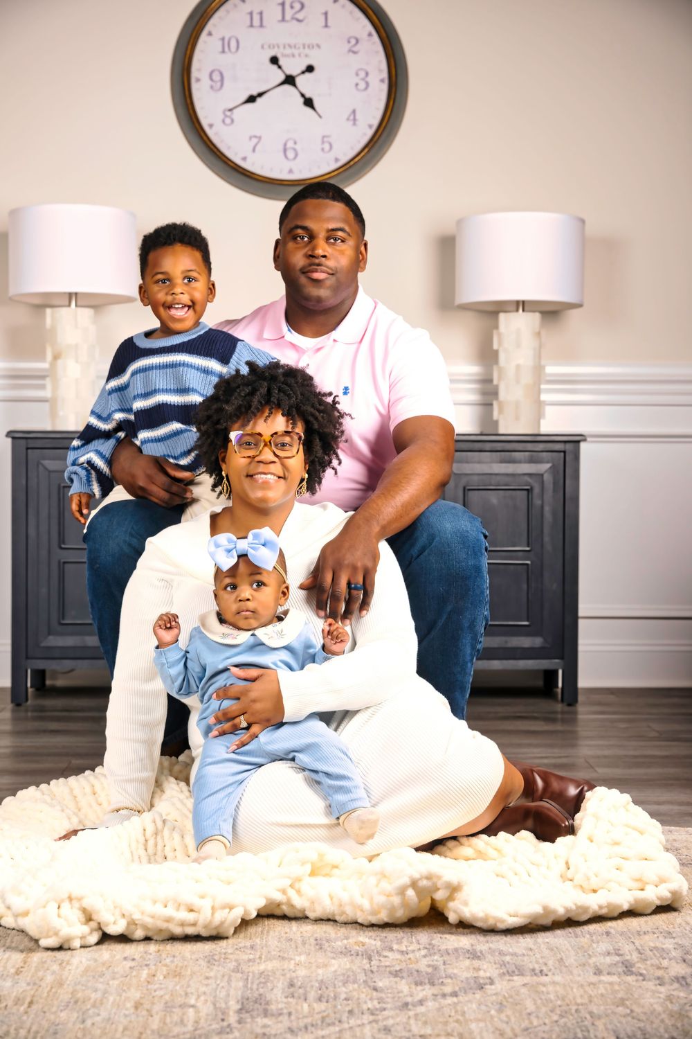 A family sits together on a cream-colored rug in a room with white walls and a wall clock.