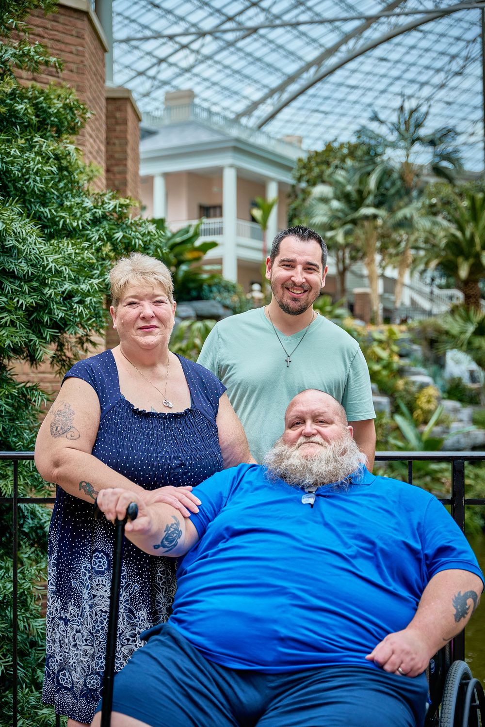 Three people posing indoors under a glass atrium, with a man in a wheelchair, a woman, and a standing man behind them.