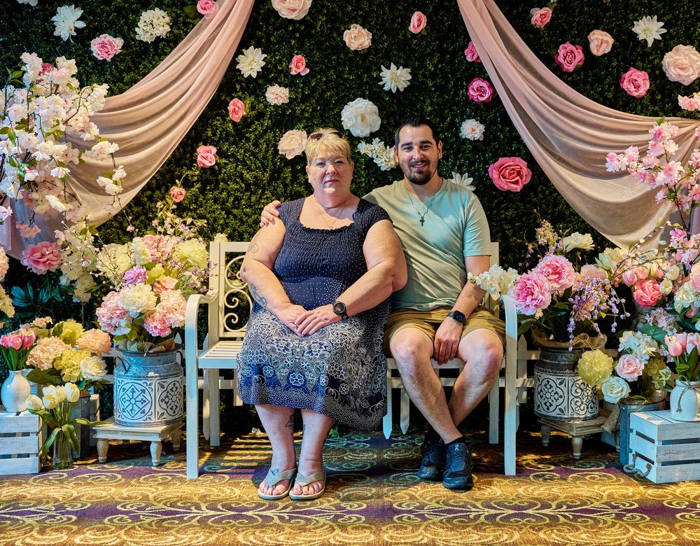 Two people sitting on a bench surrounded by colorful floral decorations and pink drapes backdrop.