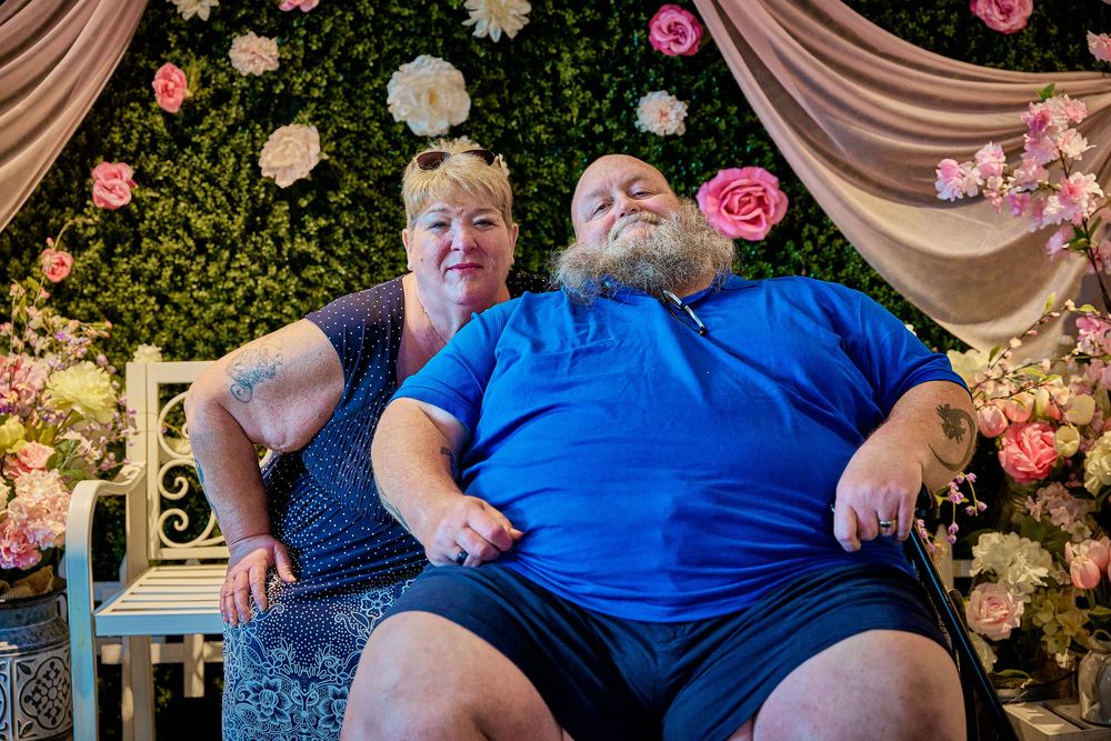 Couple posing together in front of floral backdrop with pink roses and draped fabric on decorative bench.