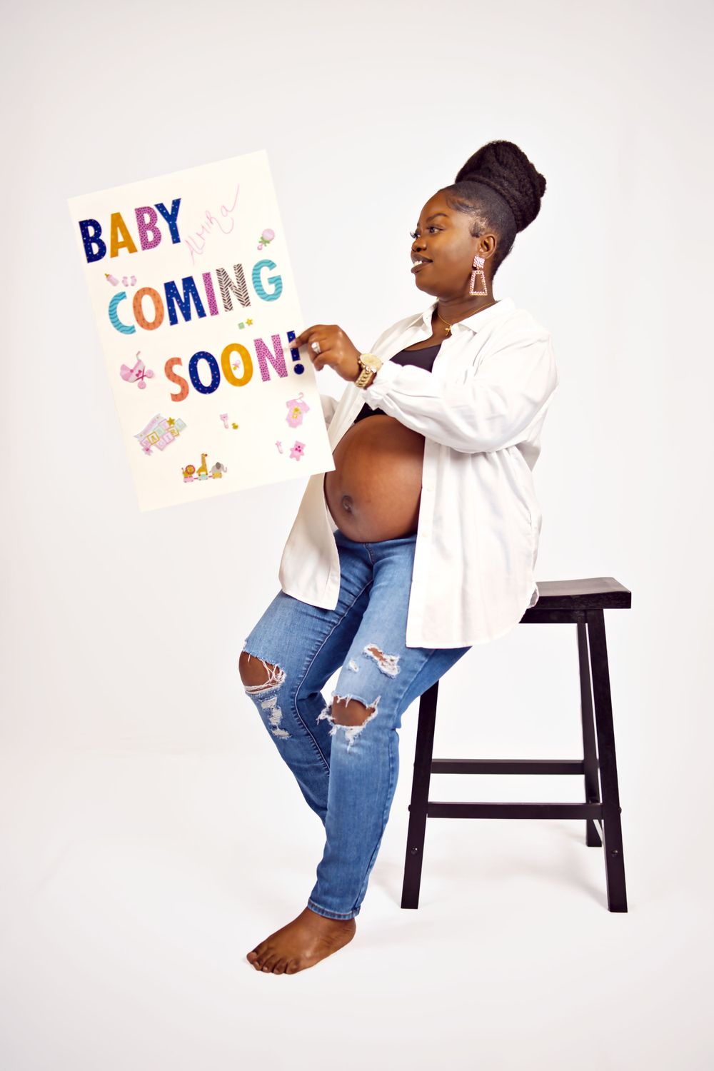 A pregnant person in ripped jeans and white shirt poses with a colorful 'Baby Coming Soon' sign against a light background.