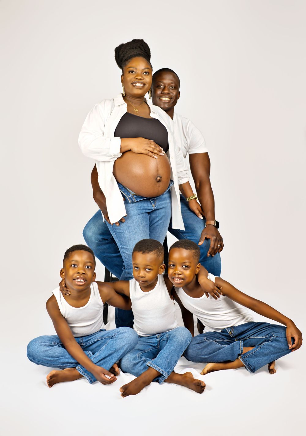 A family in matching white and denim poses together against a light background, with a pregnant mom and children seated.