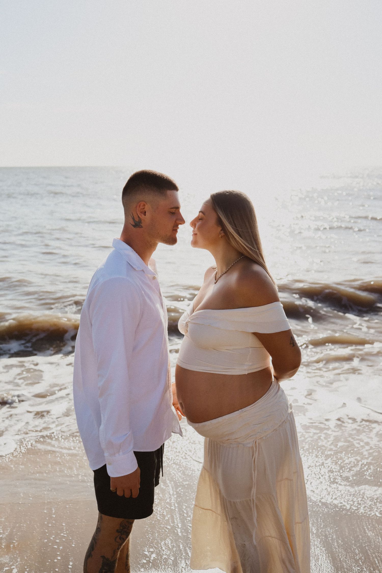 An intimate beach photoshoot of expecting couple in white attire against ocean backdrop.