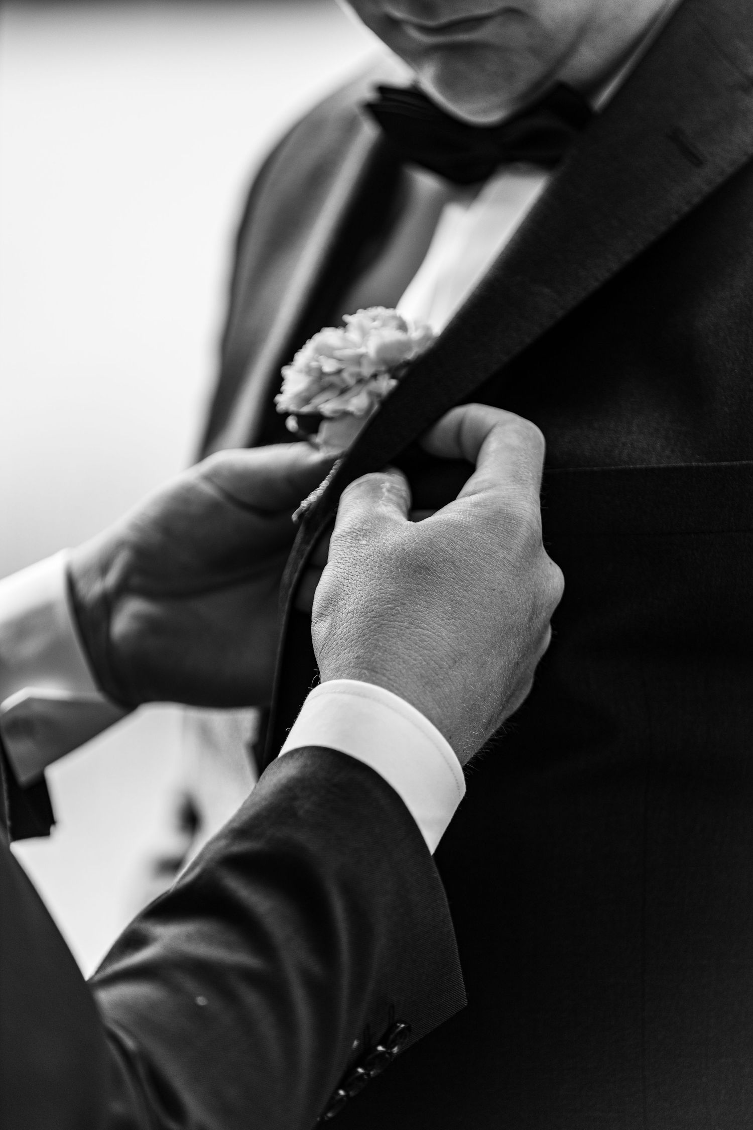 Black and white close-up of boutonniere being pinned onto groom's tuxedo lapel.