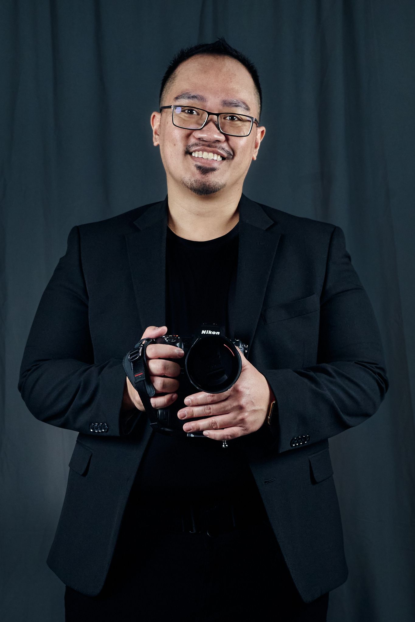 Professional photographer in black blazer and shirt smiles while holding DSLR camera against dark background.