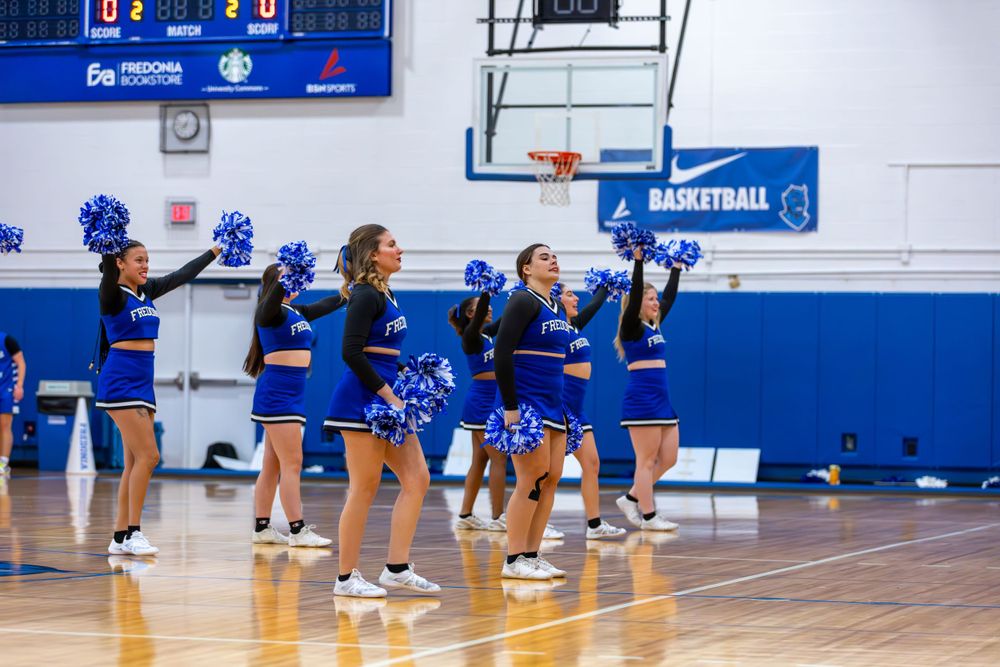 Sports team performing in a blue and white gymnasium with scoreboard visible.