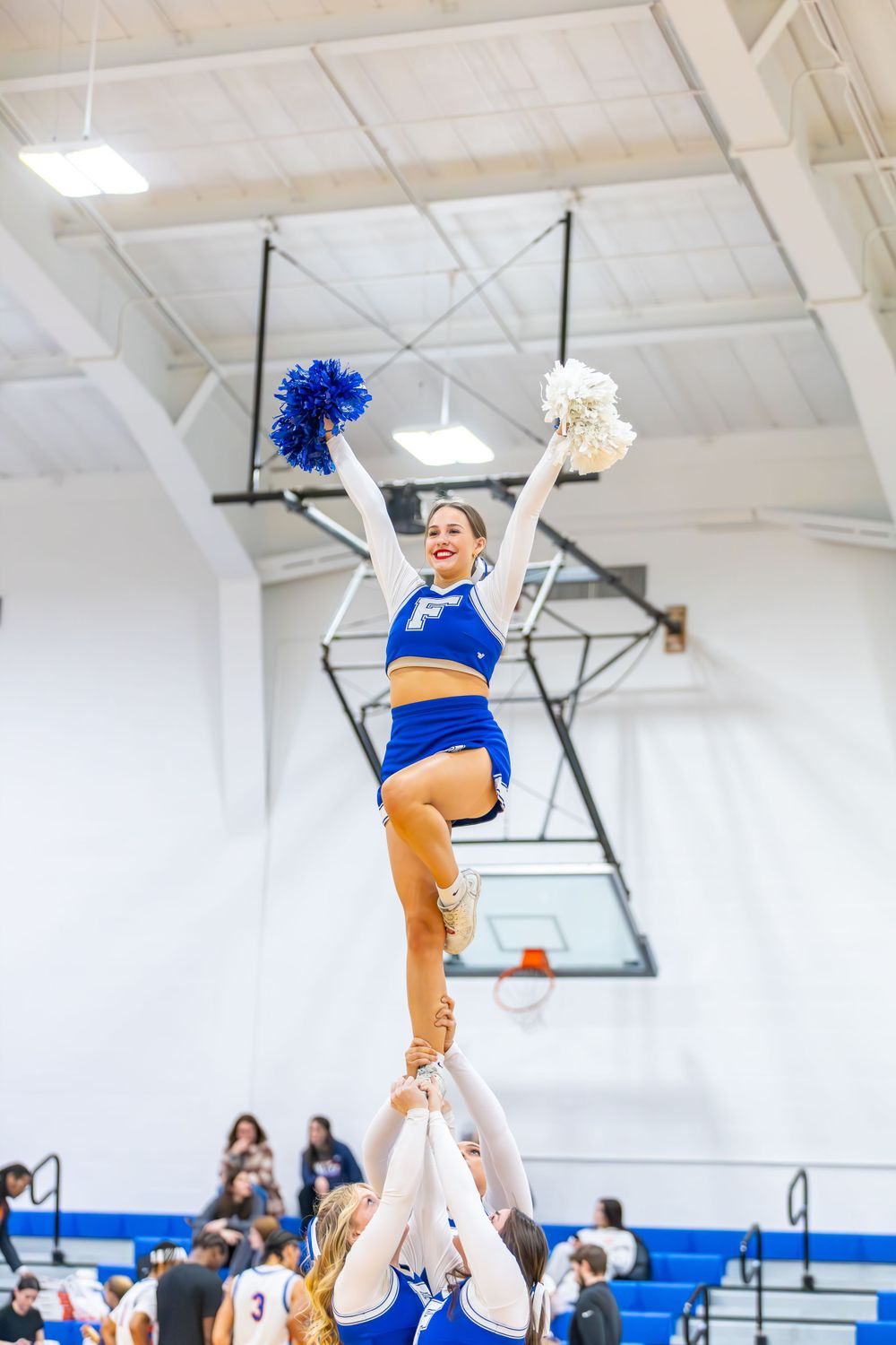 Cheerleader performs elevated stunt with blue and white pom poms in gymnasium.