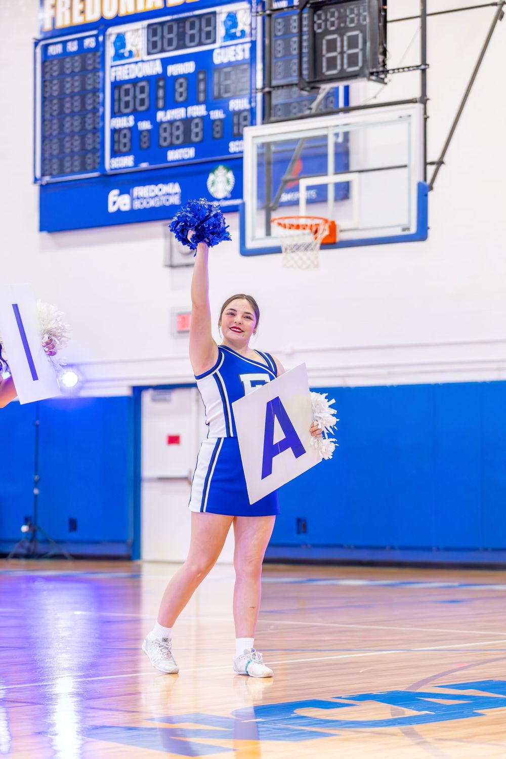 Cheerleader performs energetic moves during a basketball game in a blue and white gymnasium.