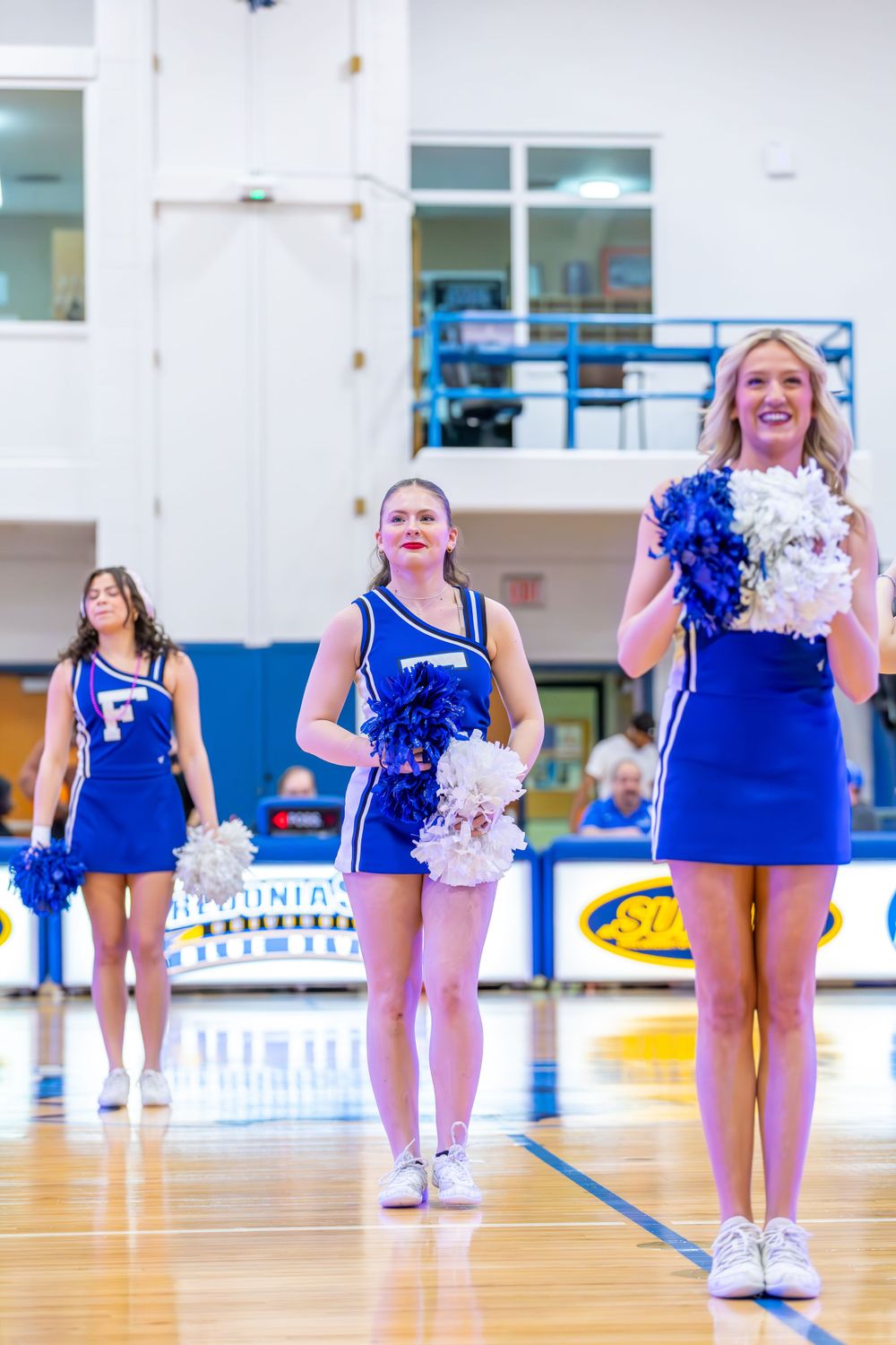 Cheerleaders in royal blue uniforms perform with pom poms on a gymnasium floor.
