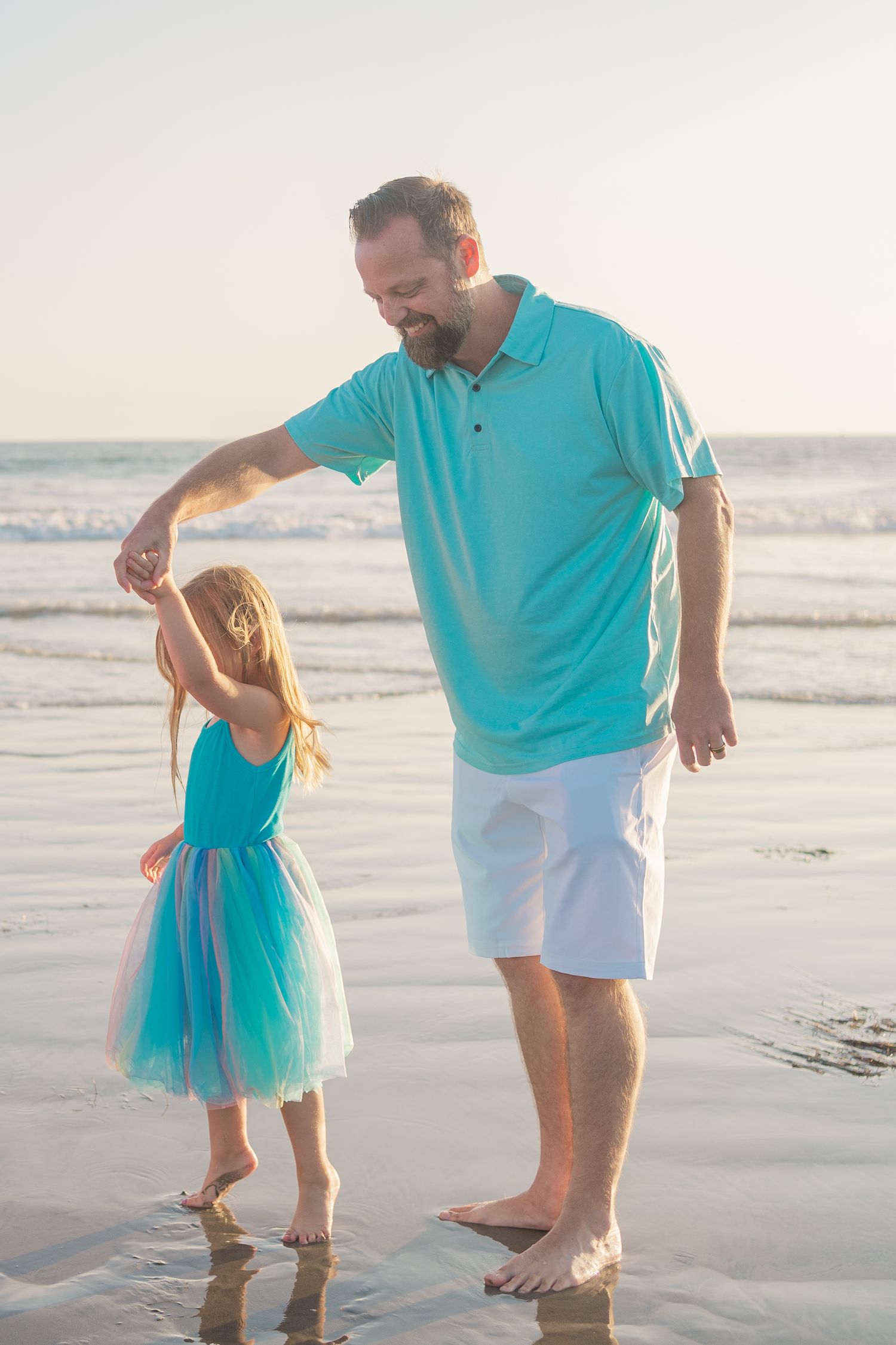Two barefoot figures in coordinating turquoise outfits hold hands while standing on a beach at sunset.