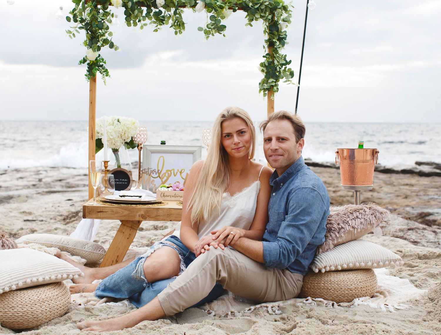 Romantic beach picnic setup with couple sitting on cushions near the ocean.