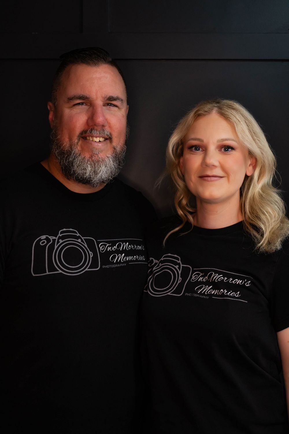 Two people wearing matching photographer t-shirts with camera logos pose against dark wall.