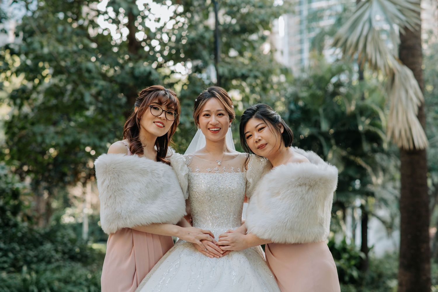 Three women in bridal attire pose together wearing white fur shawls and pink dresses.