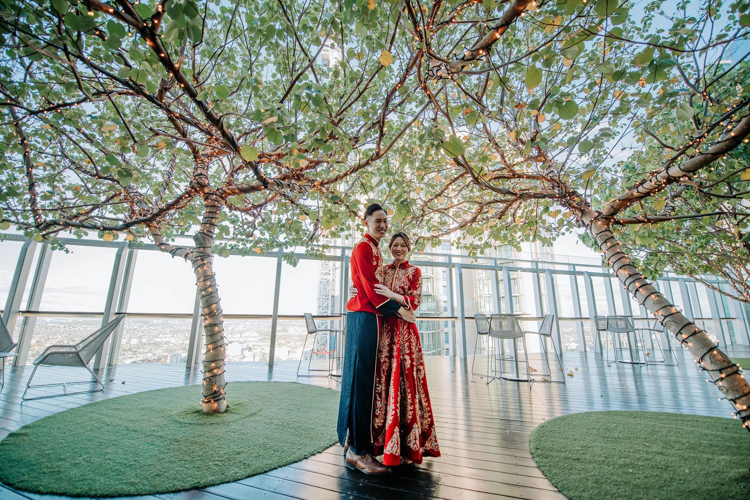 A couple in traditional wedding attire embraces under decorative trees in a modern glass-walled venue.