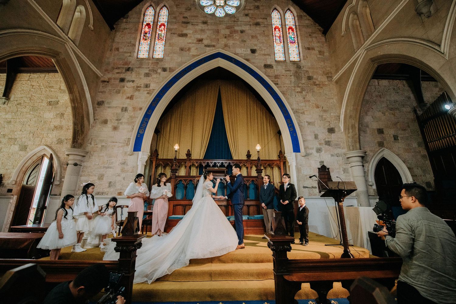A wedding ceremony takes place in a grand Gothic church with stained glass windows and vaulted arches.