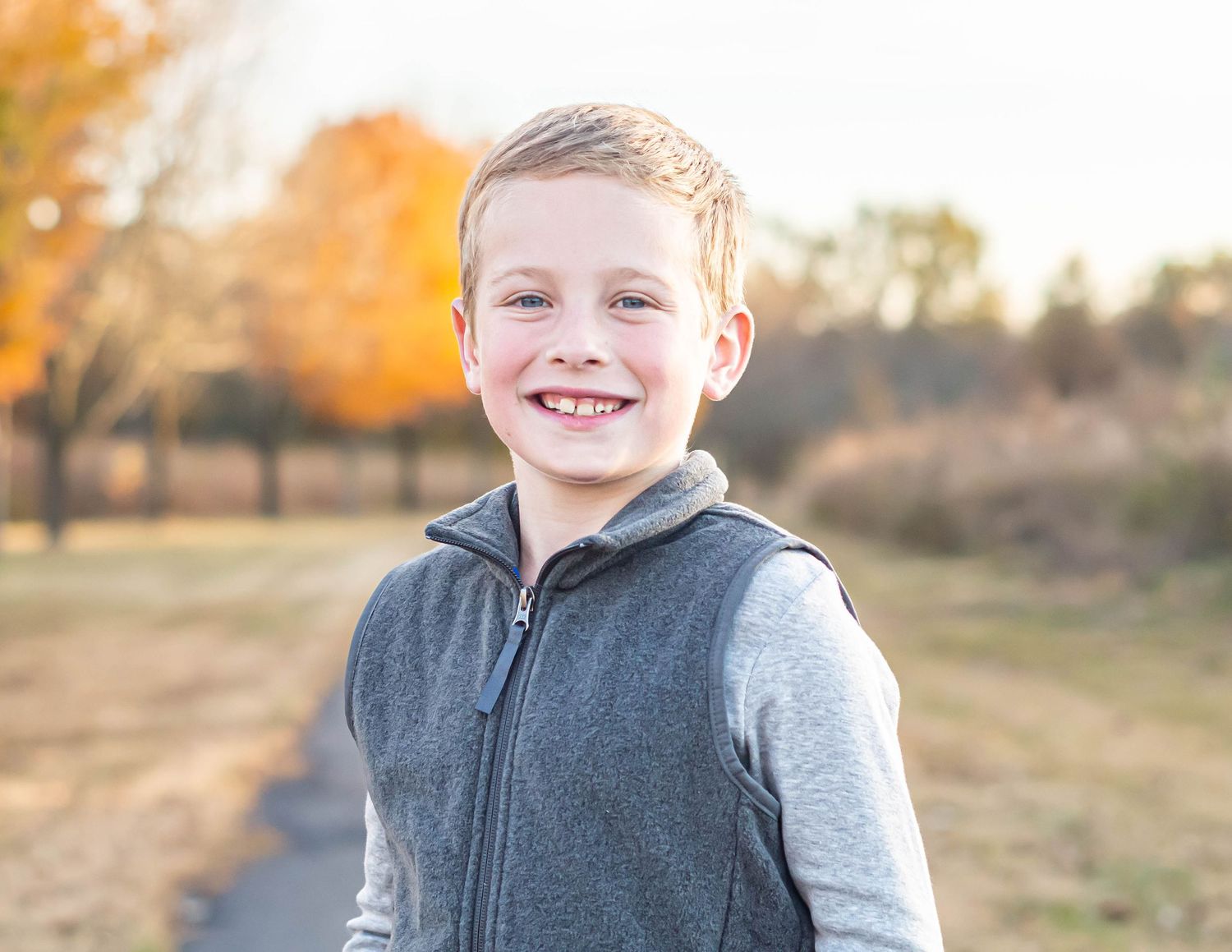 Young boy in grey vest smiling outdoors during golden hour.
