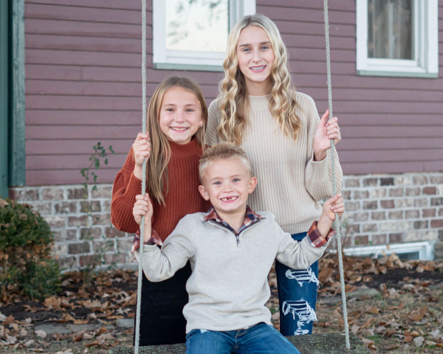 Three people pose together on a swing set against a brown house exterior.