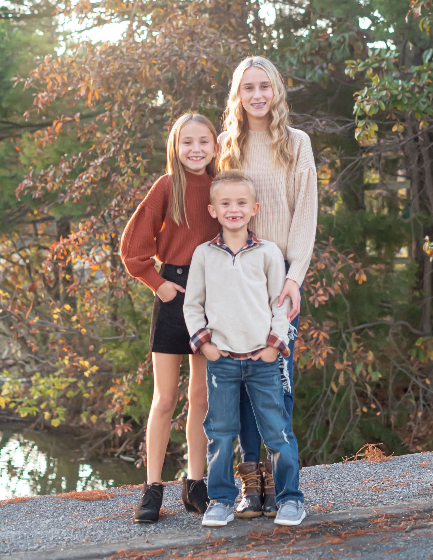 Three siblings pose together on an autumn day by a lake with fall foliage in the background.